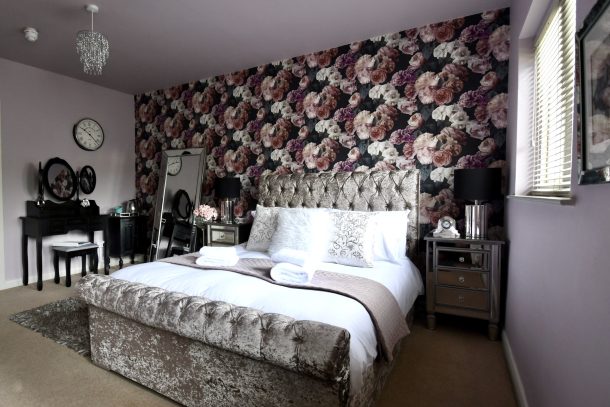 A bedroom with a floral patterned accent wall, tufted upholstered bed, mirrored nightstands, and a vanity table