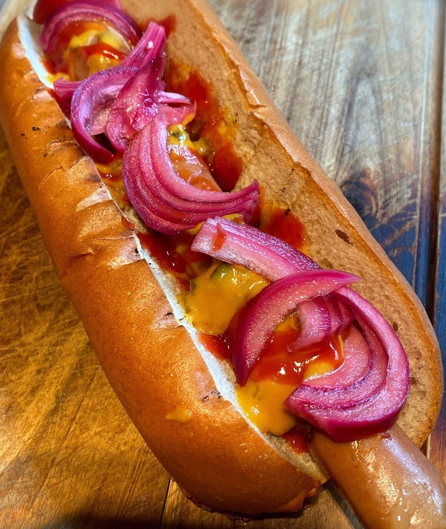 Close-up of a hot dog in a bun topped with mustard, red sauce, and pickled red onion slices, on a wooden surface