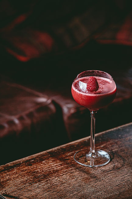A red cocktail garnished with a raspberry in a stemmed glass on a dark wooden table